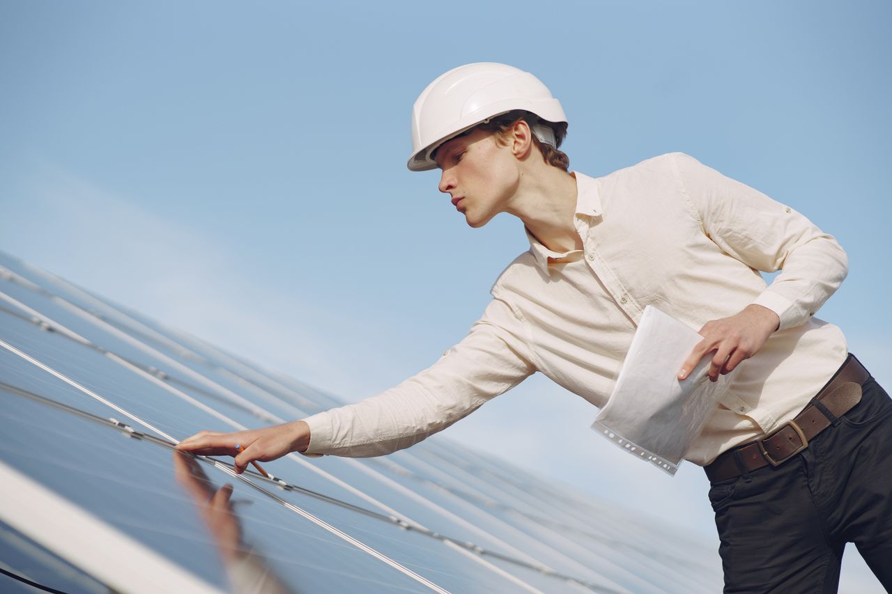 Services Low angle of young male inspector with documents in formal wear and hardhat standing against solar panels and checking details of construction