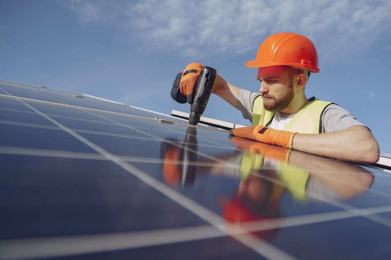 Home Low angle of serious bearded male electrician in hardhat and protective gloves installing solar photovoltaic panel system using drill
