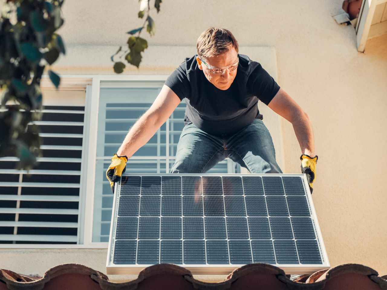 Services A man in safety glasses installs a solar panel on a house roof, promoting renewable energy.